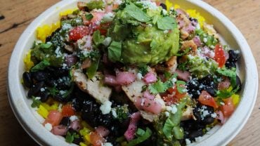 A burrito bowl with chicken, black beans, rice, pico de gallo, cotija and a scoop of guacamole on top.
