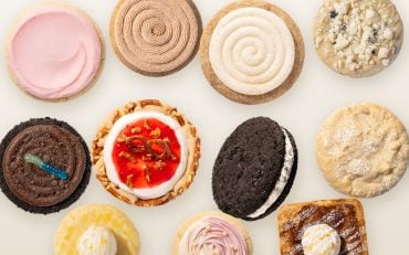 Overhead flat lay of eleven Crumbl cookies on a parchment surface, including Pink Sugar, Churro, Snickerdoodle Cupcake, Dirt Cake and Strawberry Pretzel Pie