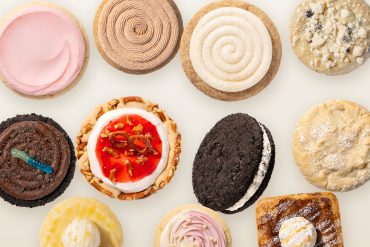 Overhead flat lay of eleven Crumbl cookies on a parchment surface, including Pink Sugar, Churro, Snickerdoodle Cupcake, Dirt Cake and Strawberry Pretzel Pie