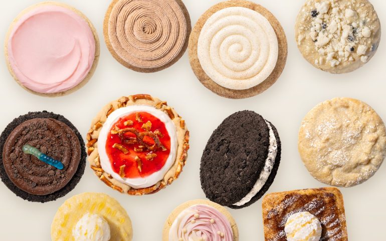 Overhead flat lay of eleven Crumbl cookies on a parchment surface, including Pink Sugar, Churro, Snickerdoodle Cupcake, Dirt Cake and Strawberry Pretzel Pie