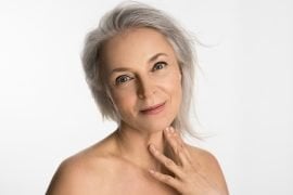 A woman in her sixties with grey hair and natural skin texture, resting a hand at her collarbone against a white background