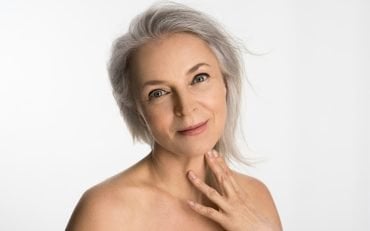 A woman in her sixties with grey hair and natural skin texture, resting a hand at her collarbone against a white background