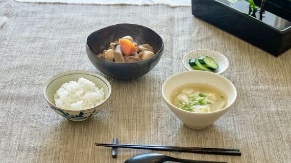 A Japanese meal laid out on a linen mat: a bowl of rice, simmered vegetables with carrot and mushroom, cucumber pickles and a bowl of miso soup with tofu.