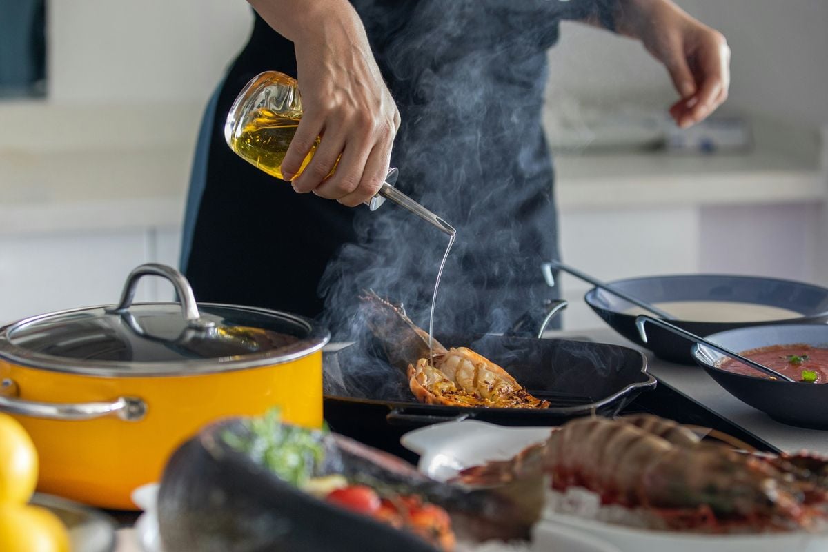 A cook pouring golden oil from a glass cruet into a smoking pan.