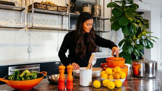 Woman in a bright kitchen lifting the lid of an orange pot, with lemons and a bowl of greens on the counter
