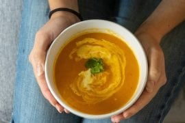 Two hands cradling a white bowl of creamy orange soup with a swirl of cream and a sprig of parsley