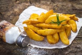 A tipped-over salt shaker spilling salt beside a pile of fried potato wedges on rustic wood